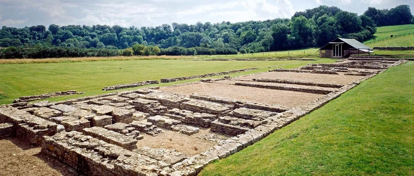 The low stone wall footings of North Leigh Roman Villa running across a lawn towards the timber mosaic house, with woodland and pasture beyond