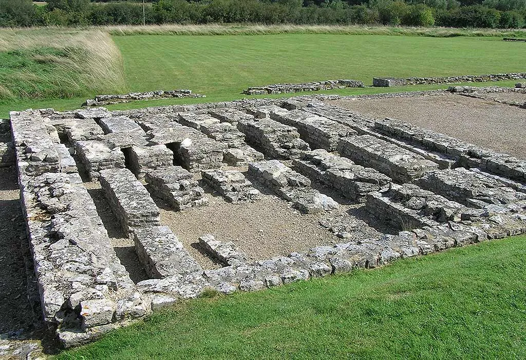 The rows of stone pillars of a hypocaust heating system at North Leigh Roman Villa, on gravel beside a mown lawn and hedgerow