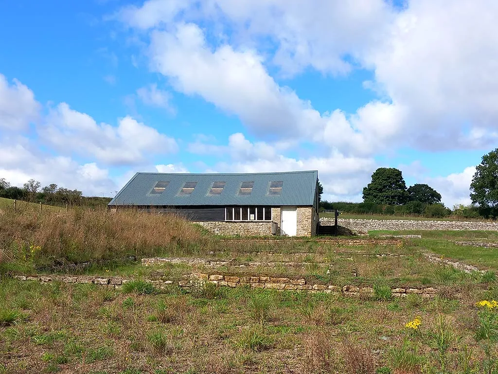 The modern cover building over the mosaic at North Leigh Roman Villa, behind low Roman wall footings in rough grass under a bright cloudy sky