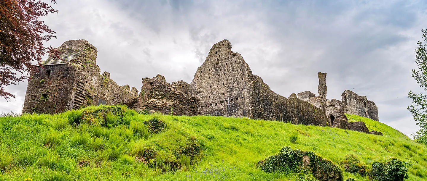 The ruined walls of Okehampton Castle rising above a grassy bank under a cloudy sky
