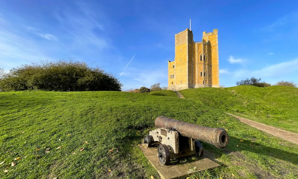 An old cannon on the grass below the keep on its mound under a deep blue sky