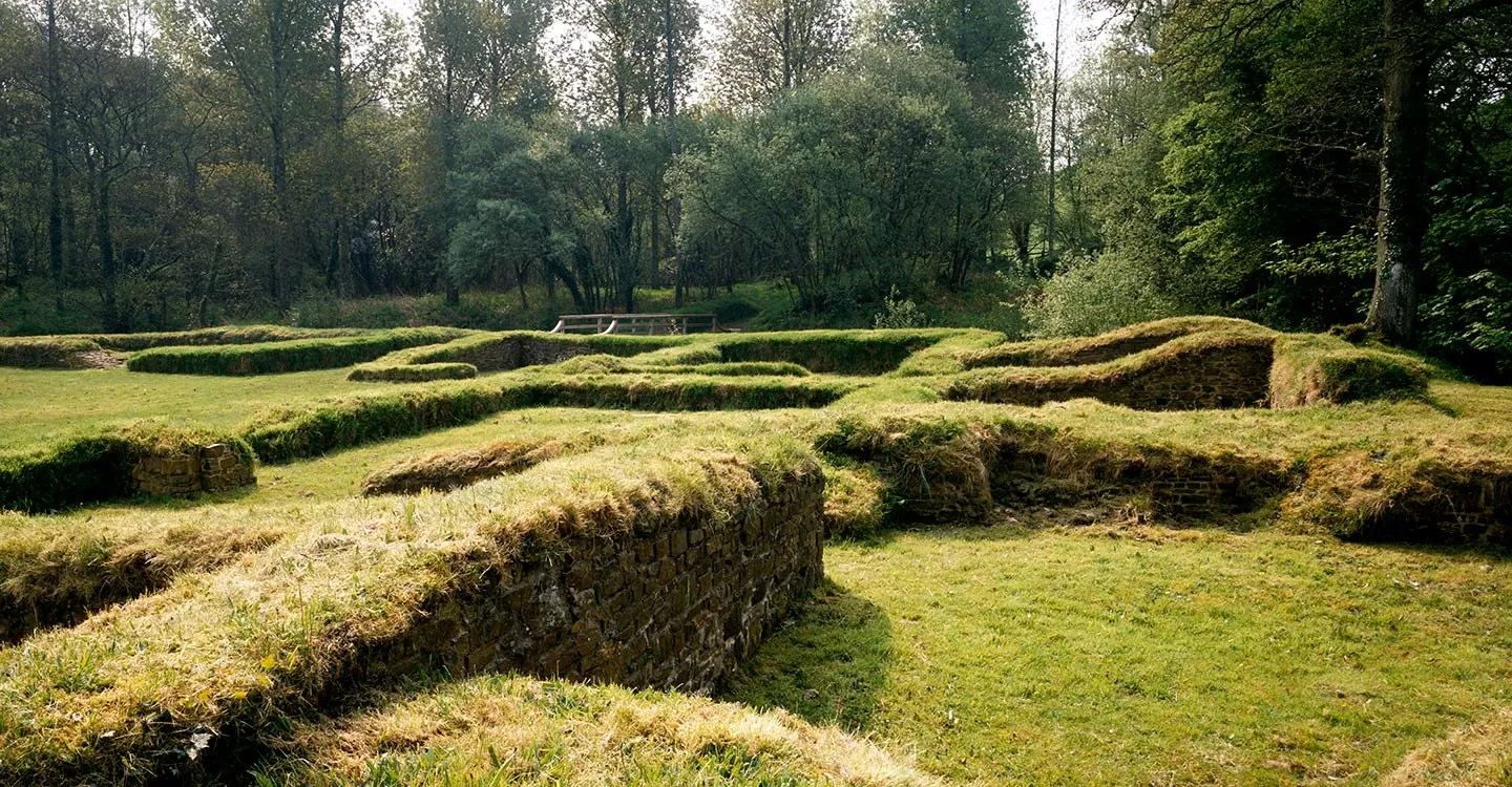 The grass-topped stone wall footings of Penhallam Manor in a sunny woodland clearing, with a wooden footbridge over the moat beyond