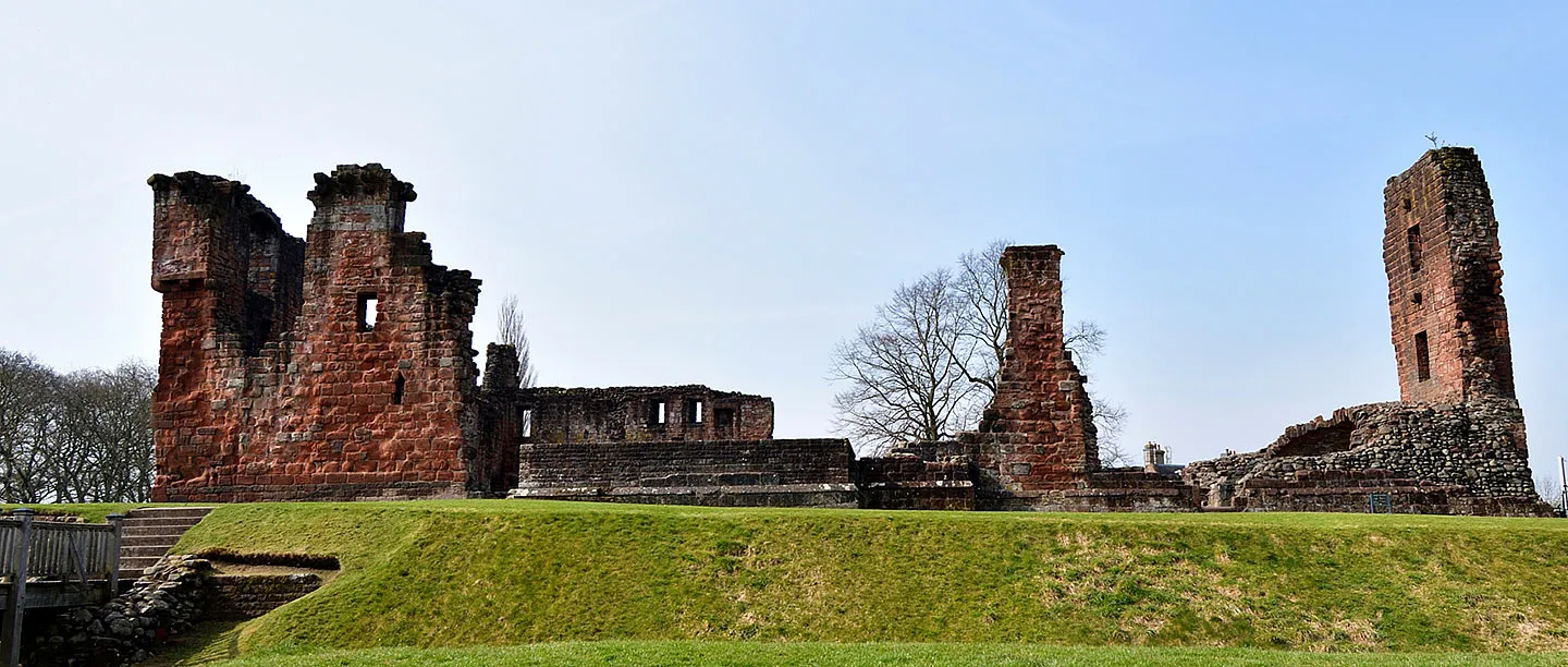 Ruined red sandstone towers of Penrith Castle above the grassy moat, with wooden steps and a bridge on the left