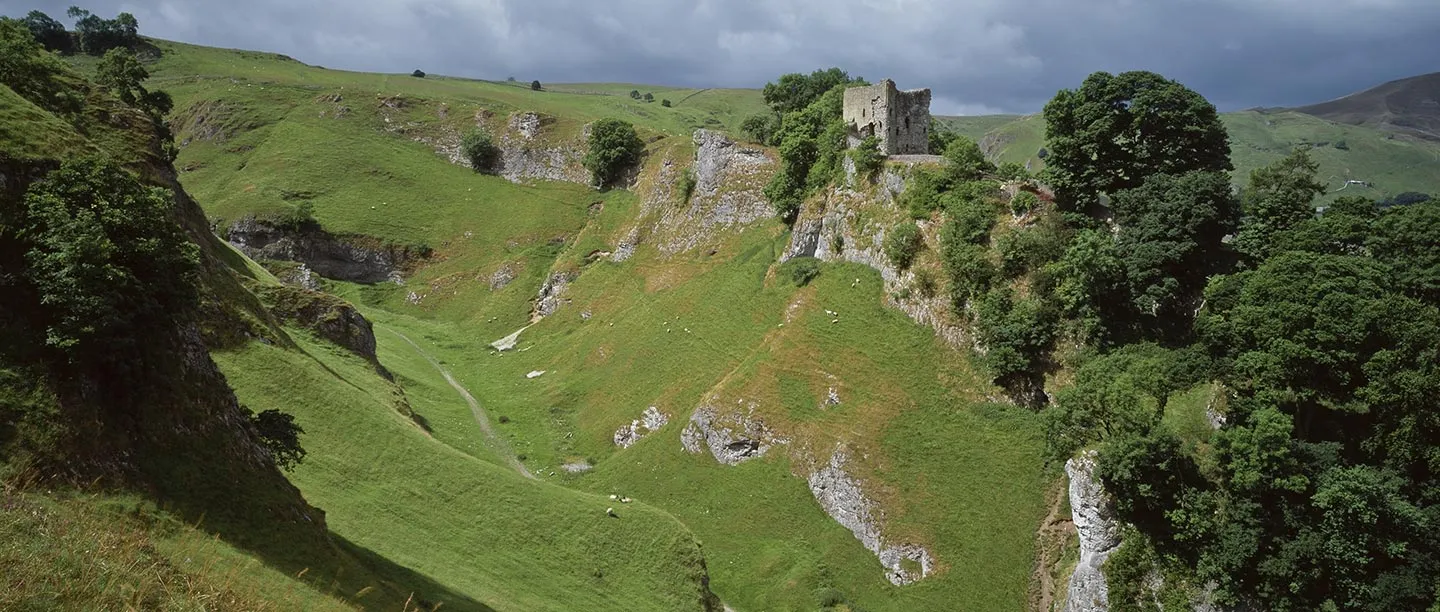 Peveril Castle's keep on its wooded crag above the green limestone gorge of Cave Dale, under a stormy sky