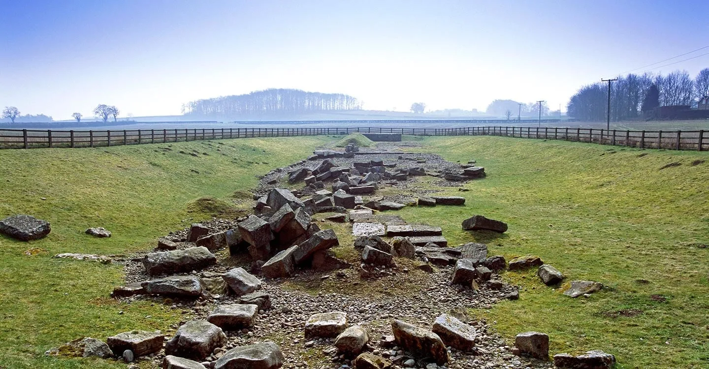 Tumbled masonry blocks of the Roman bridge at Piercebridge in a grassy hollow, with misty fields and woods beyond