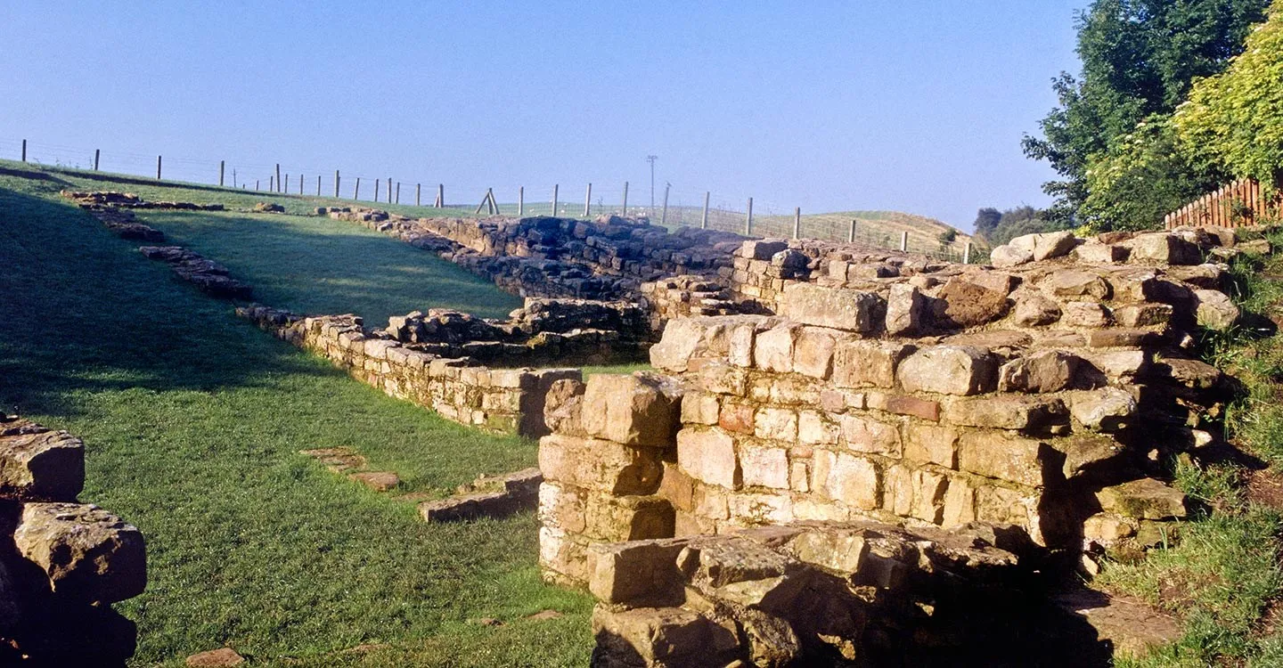 The stone walls of Poltross Burn milecastle in low morning sun, with a fence line running up the hillside behind