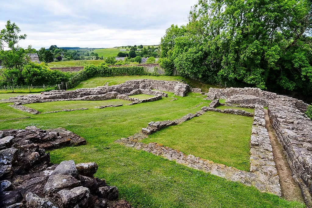 Poltross Burn milecastle from above, its low walls and grassy rooms with trees, a railway line and green hills beyond