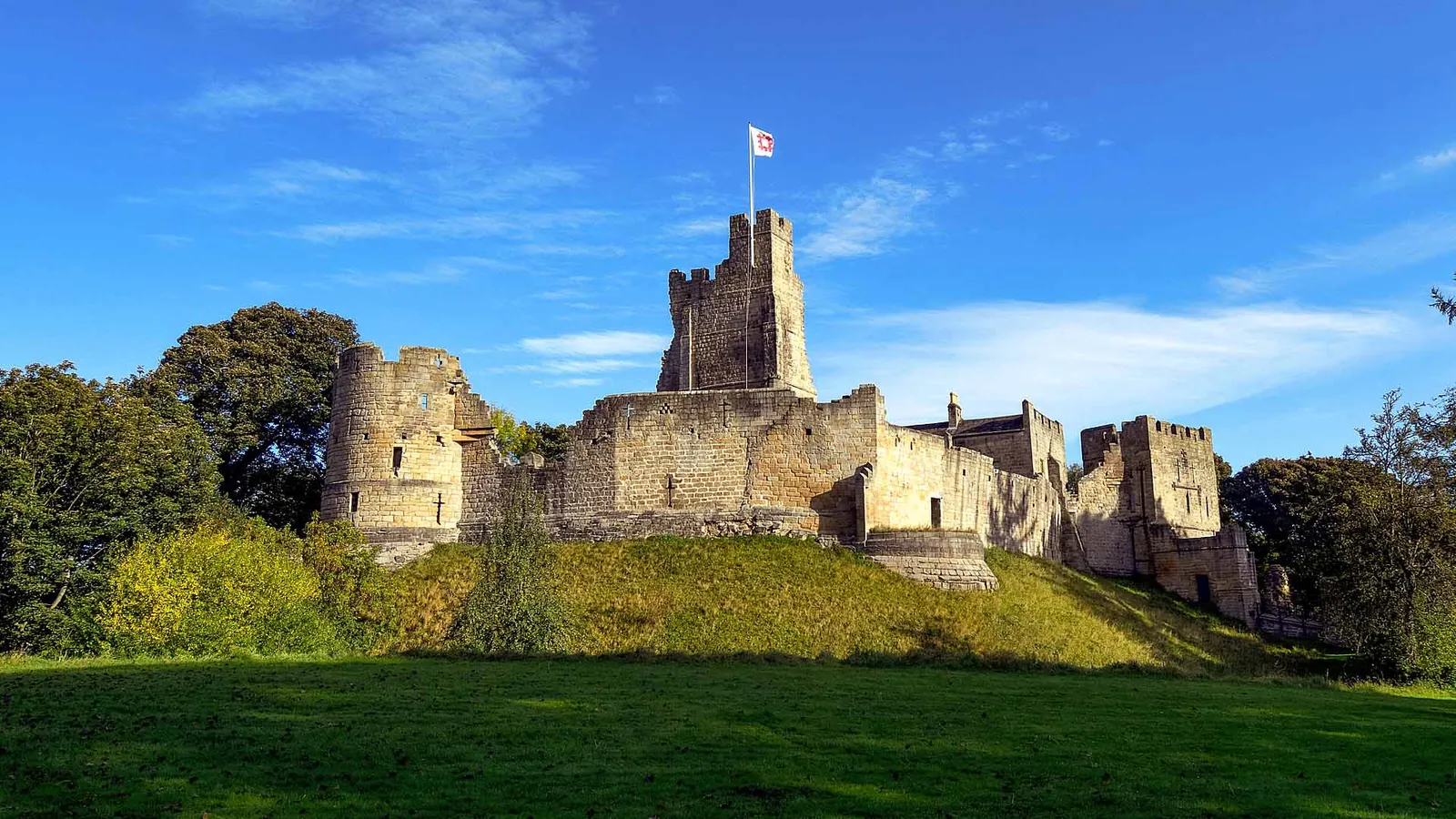 Prudhoe Castle's keep, round tower and battlemented curtain wall above a grassy bank, with an English Heritage flag against a blue sky