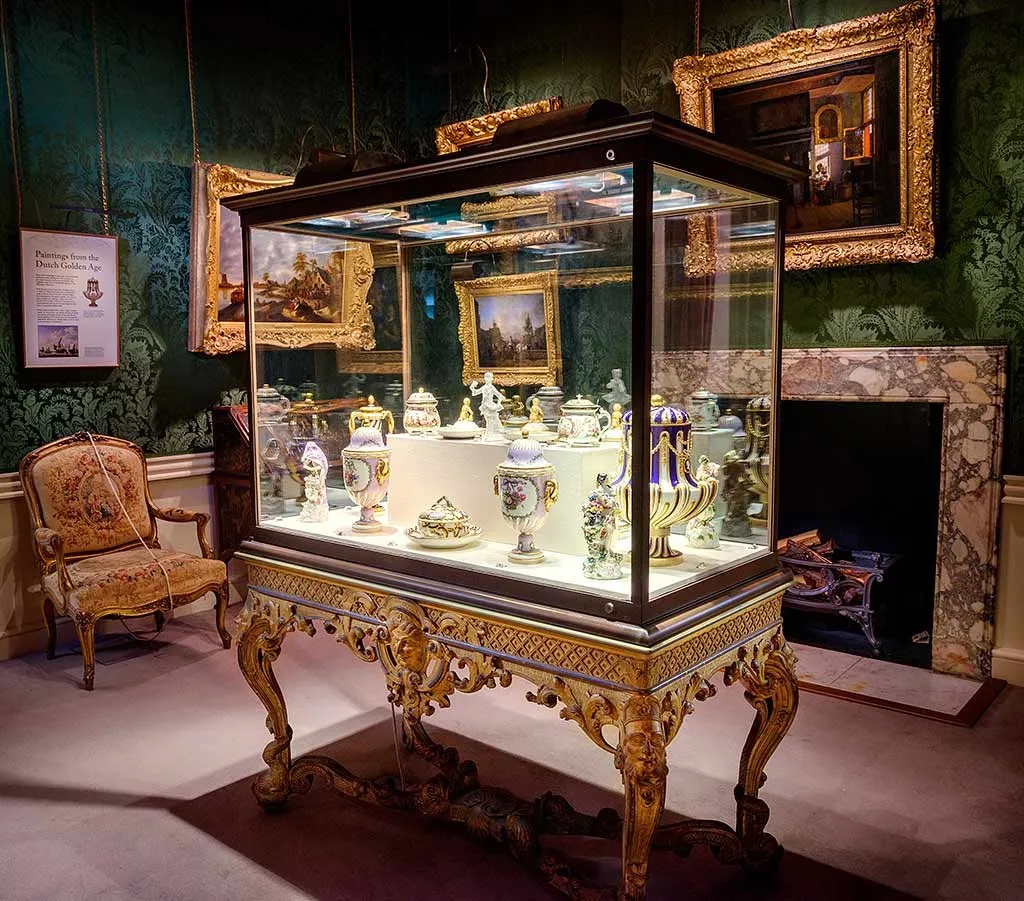 A glass case of Sevres and Meissen porcelain on a carved gilt table in the green silk room at Ranger's House, beside paintings and a marble fireplace
