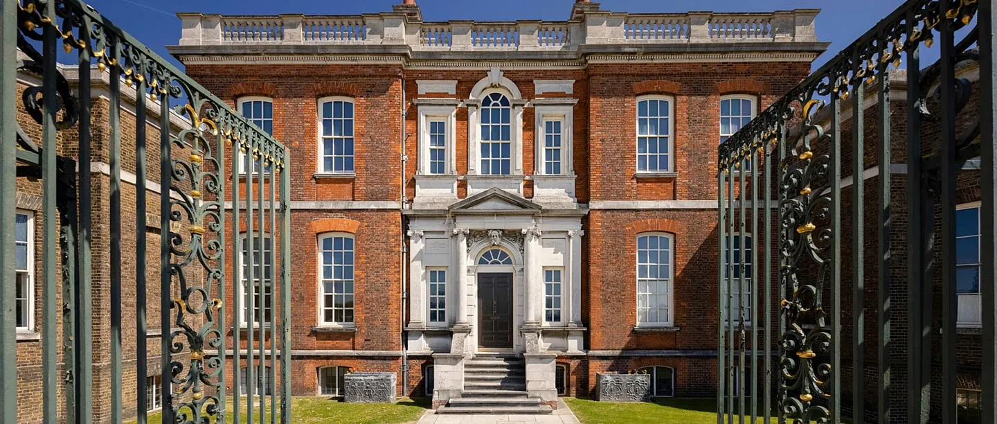 The red-brick facade and white stone doorcase of Ranger's House, framed by open wrought-iron gates with gilded scrollwork under a blue sky