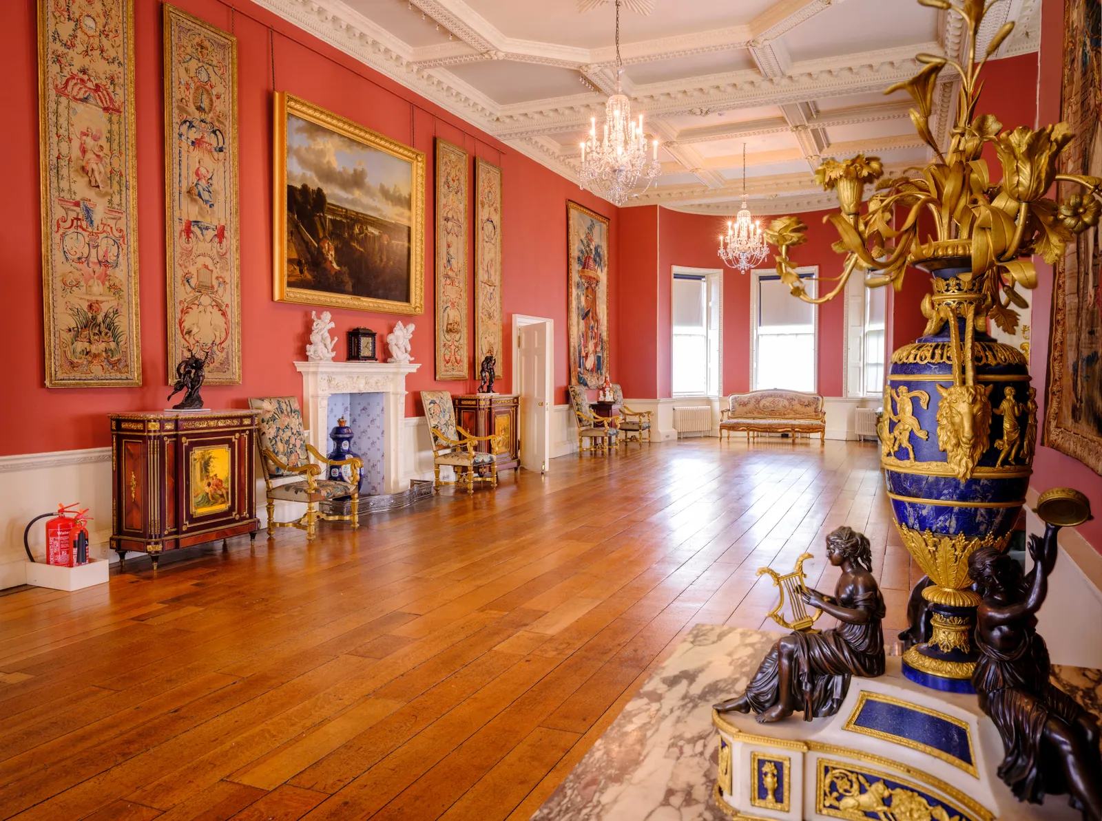 The long gallery at Ranger's House with red walls, tapestries, a landscape painting, crystal chandeliers and a gilt and lapis lazuli vase