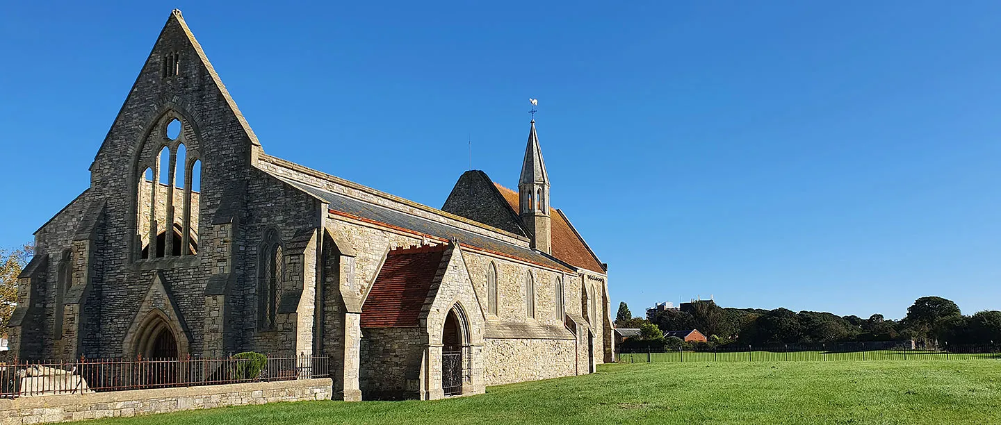 The Royal Garrison Church in Portsmouth, a roofless Gothic nave with an empty tracery window beside the roofed chancel and bellcote, on a lawn under a deep blue sky