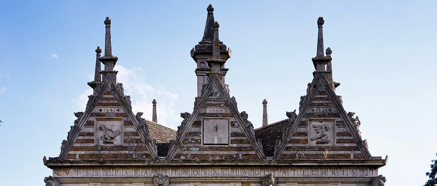 The three carved gables of Rushton Triangular Lodge with pinnacles, a dove, a pelican and a sundial against a blue sky