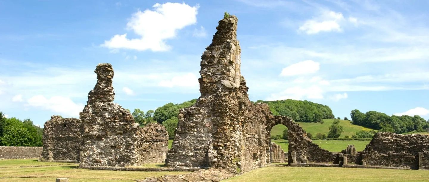 The jagged ruined walls and a round arch of Sawley Abbey on a mown lawn, with sheep fields and a wooded hill beyond under a blue sky