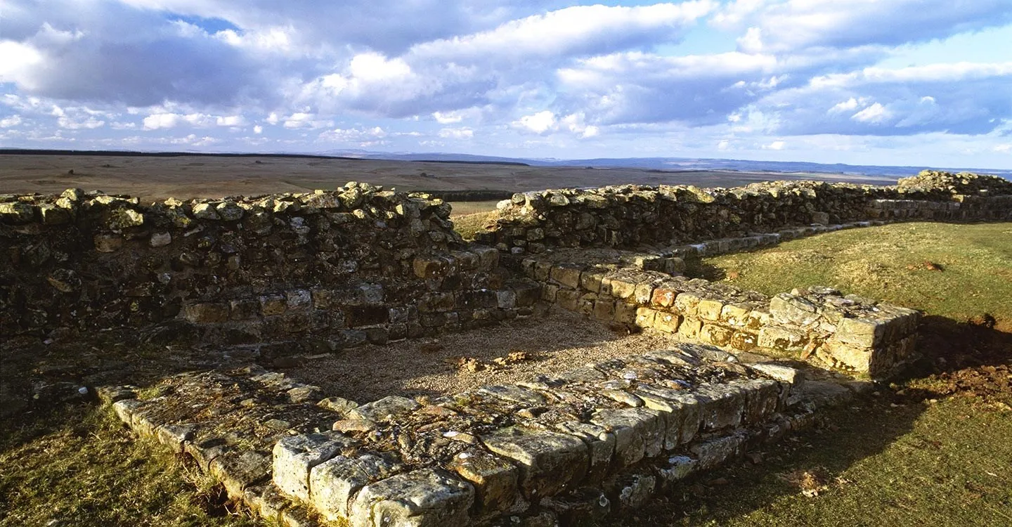The stone walls of a milecastle on Hadrian's Wall at Sewingshields, lit by low sun, with open moorland and a wide cloudy sky beyond