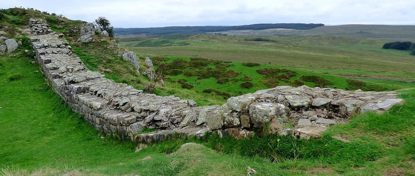 Hadrian's Wall curving along the top of Sewingshields Crags, with rocks, green fields and dark forest stretching away to the north
