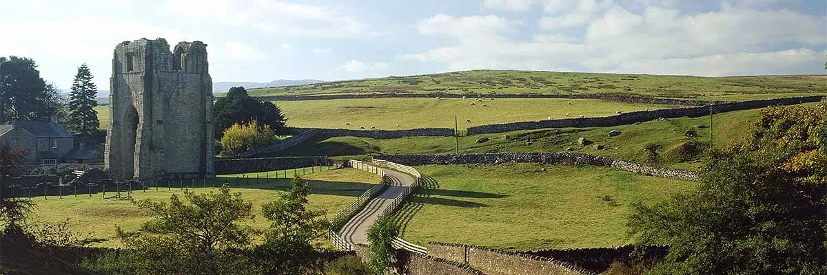 The tall ruined west tower of Shap Abbey beside a farmhouse, with a fenced lane winding past dry-stone walls into fell pasture dotted with sheep