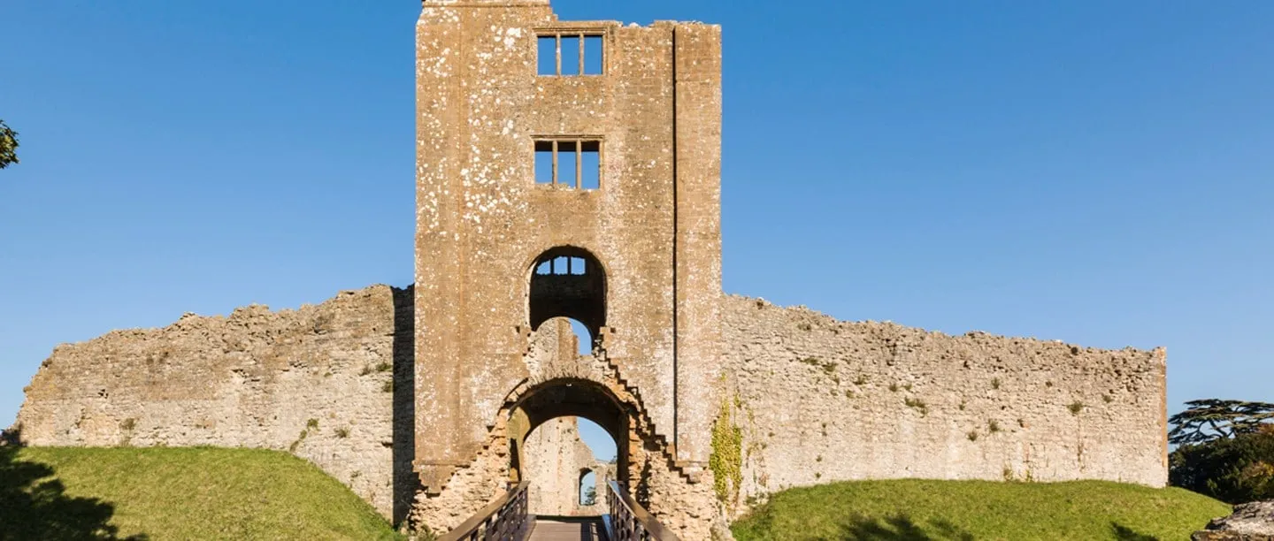 The tall ruined gatehouse of Sherborne Old Castle with its arched entrance and a footbridge, beside the curtain wall under a clear blue sky