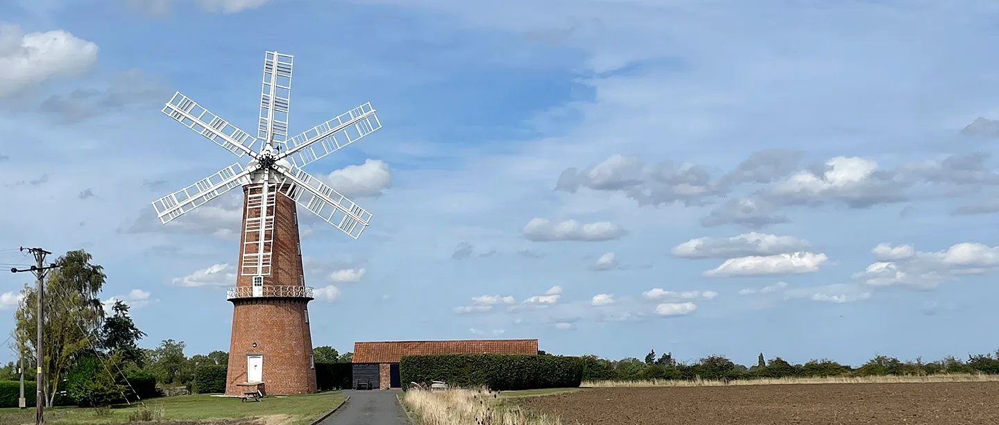 Sibsey Trader Windmill, a tall red-brick tower mill with six white sails, beside a barn and ploughed fields under a blue sky