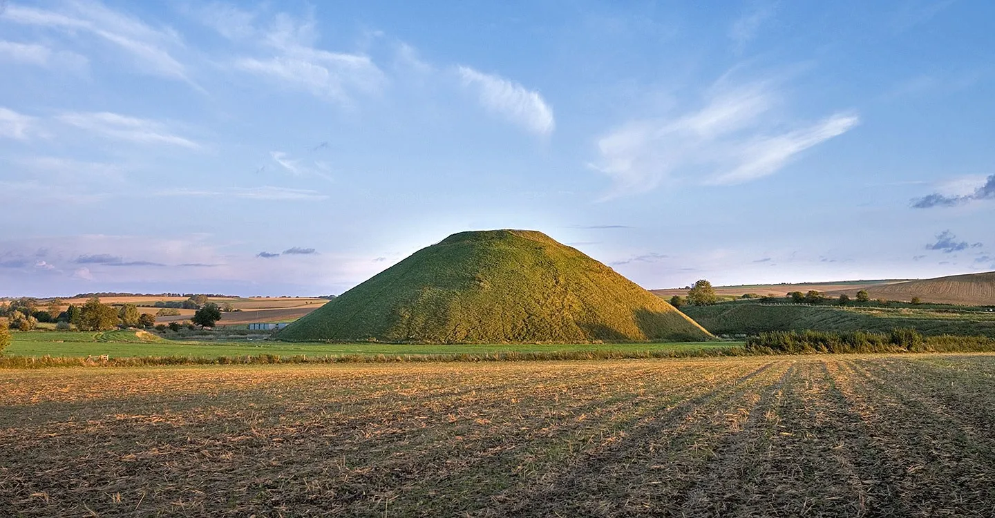 Silbury Hill, a huge grassy prehistoric mound, rising beyond a stubble field and hedgerow in warm evening light under wispy clouds