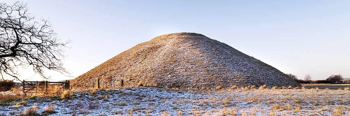 Silbury Hill dusted with frost and snow in low winter sun, behind a wooden gate and a bare tree