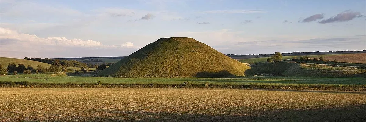 Silbury Hill in evening sunlight across a ploughed field, with rolling Wiltshire downs and trees beyond