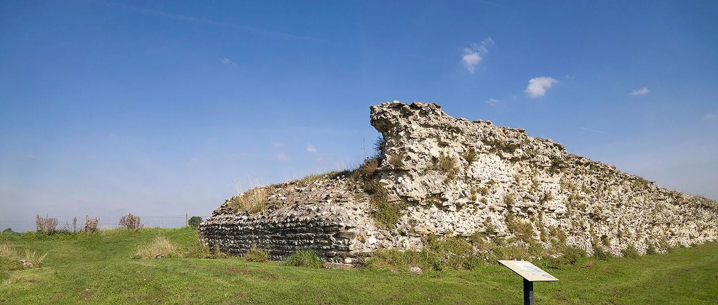 A long stretch of Silchester's Roman flint town wall beside grass and an interpretation panel, under a blue sky
