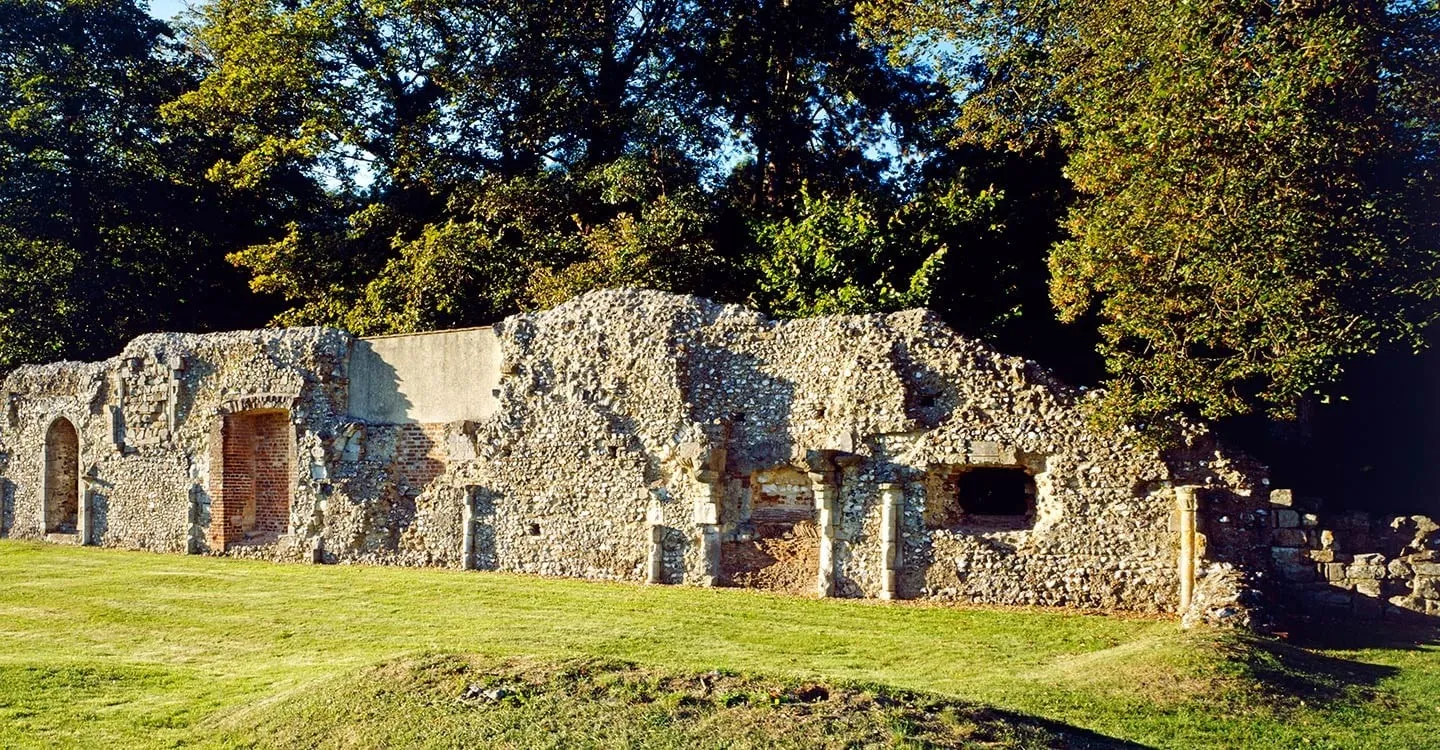 The ruined flint refectory wall of Southwick Priory with a brick-lined doorway and window openings, lit by low sun under tall trees