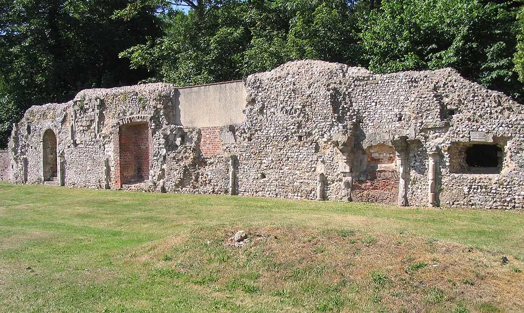 The long flint and brick wall of Southwick Priory's refectory beside a mown lawn and a grassy mound, under dense summer trees