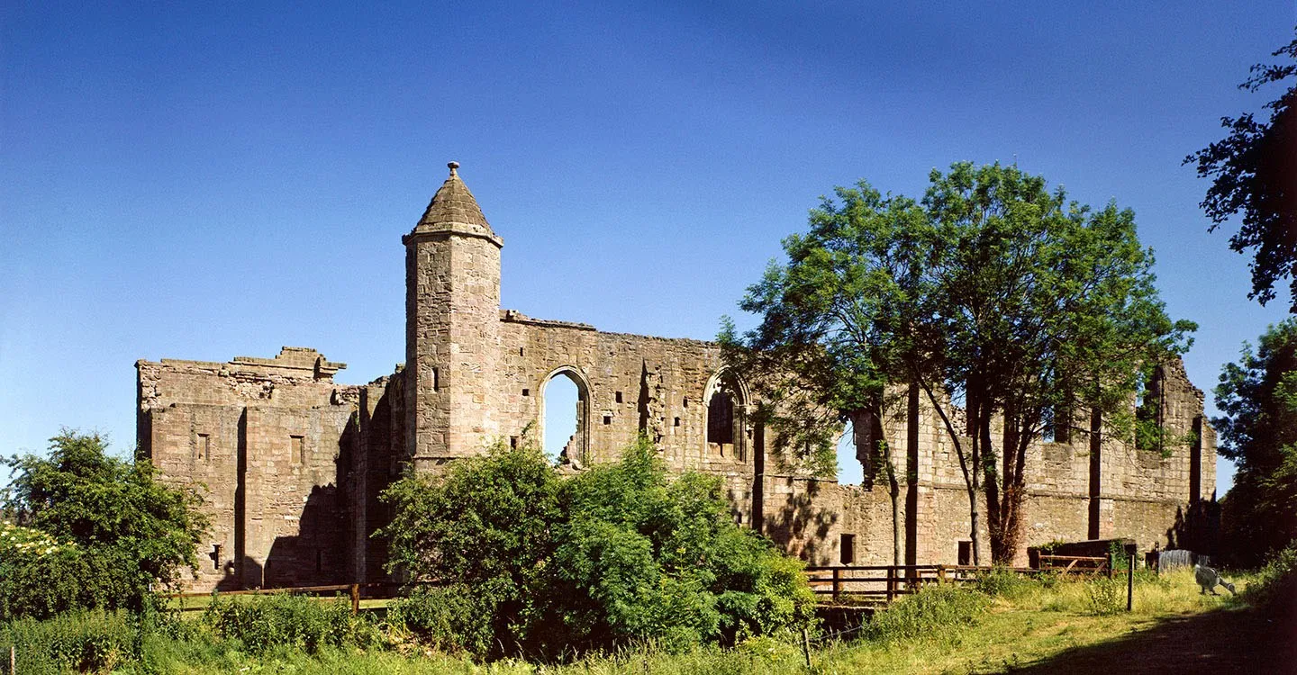 The ruined sandstone hall and conical-roofed stair turret of Spofforth Castle behind trees under a deep blue summer sky