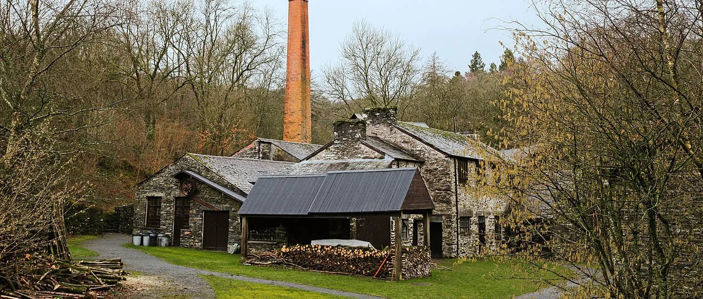 The slate-stone buildings of Stott Park Bobbin Mill with a tall brick chimney, a log store and a gravel path, among bare woodland trees