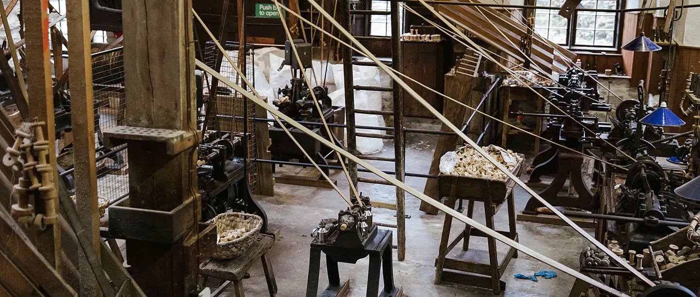 Rows of bobbin-making lathes driven by long belts from overhead shafts in the Stott Park mill, with baskets of bobbins and a window beyond