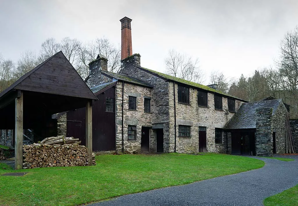 The two-storey stone mill building at Stott Park with a brick chimney, an open-sided timber shed stacked with logs and a curving path across a lawn