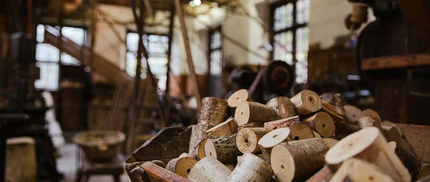 A basket of cut birch blanks with holes bored through them in the Stott Park workshop, with machinery and windows softly blurred behind