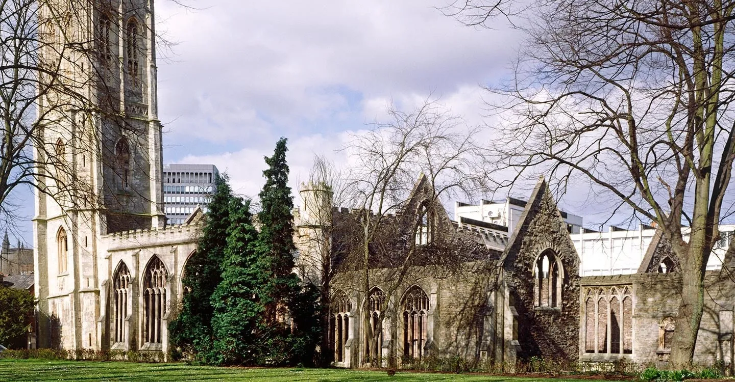 The tall leaning tower and roofless Gothic nave of Temple Church in Bristol, bombed in 1940, beside a lawn under spring trees