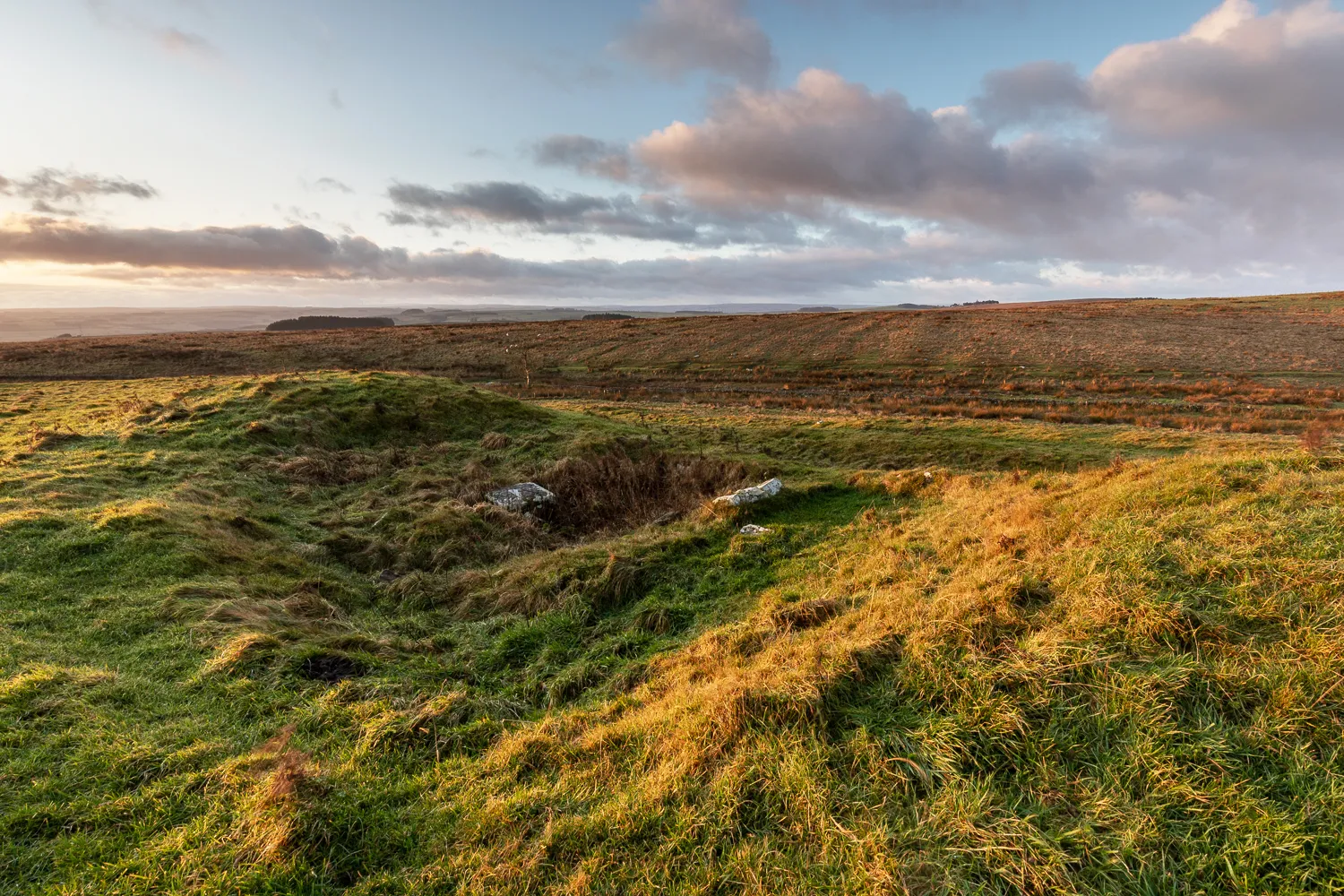 Grassy earthworks of the Roman fort at Carrawburgh catching golden evening light, with moorland stretching away under pink clouds