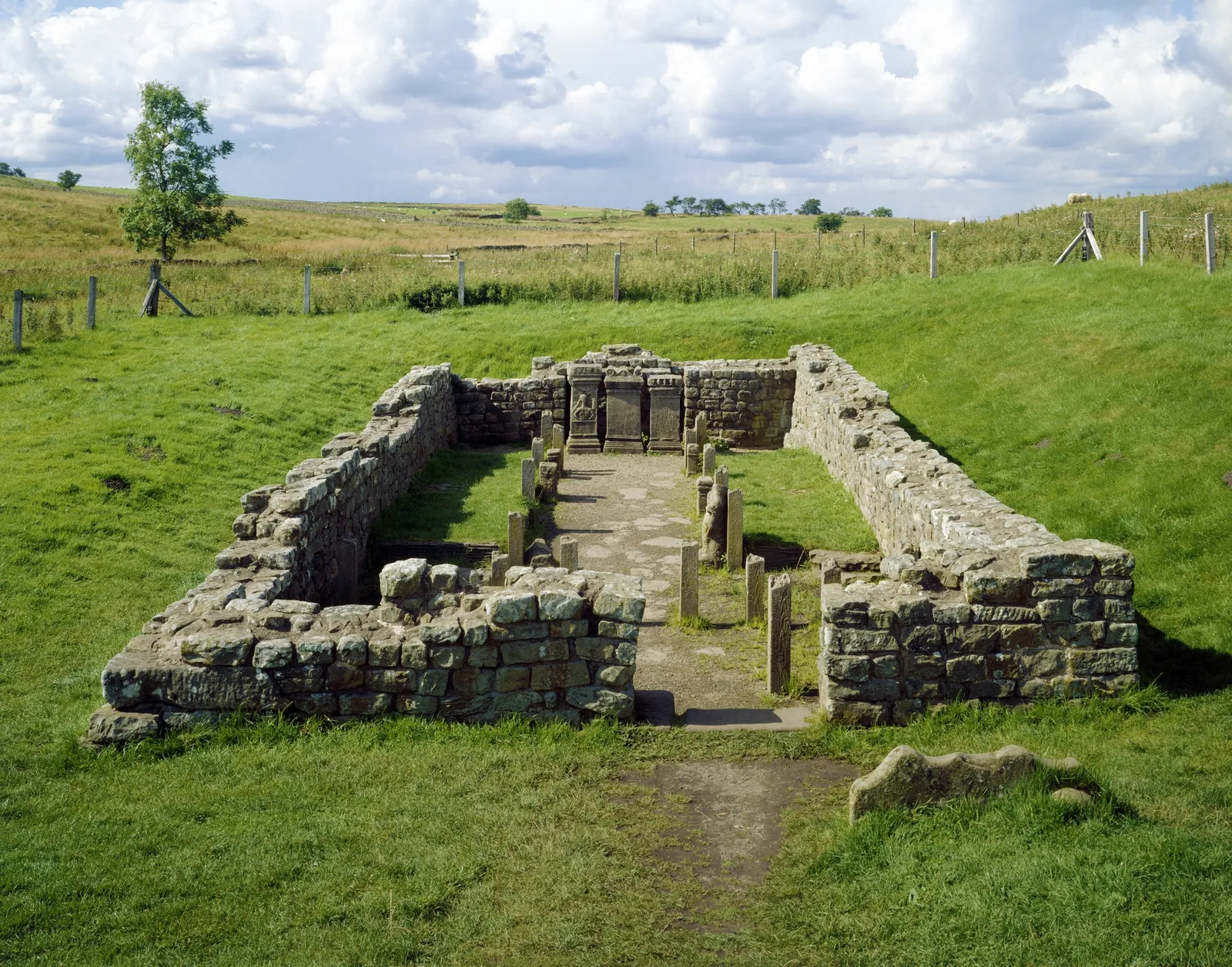 The low stone walls of the Temple of Mithras at Carrawburgh, with three altars at the far end, set in green pasture under a big cloudy sky