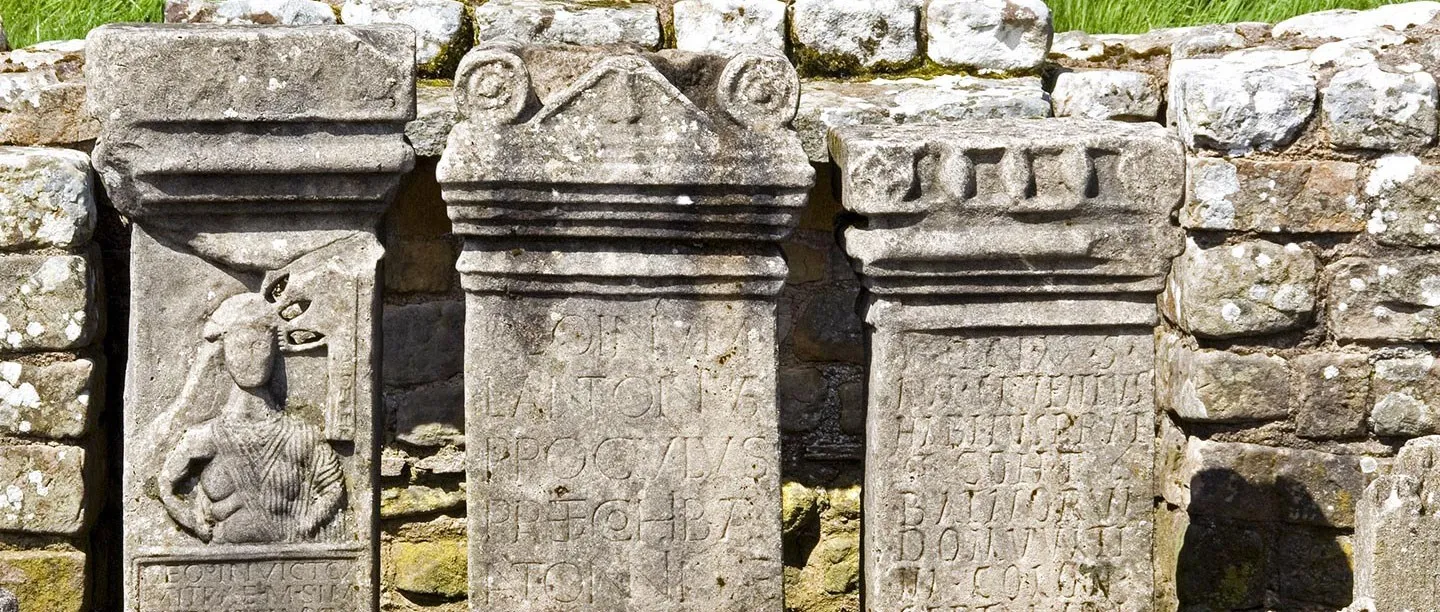 Three Roman altars in the shrine of the Temple of Mithras at Carrawburgh, one carved with the god's radiate bust, against a dry-stone wall