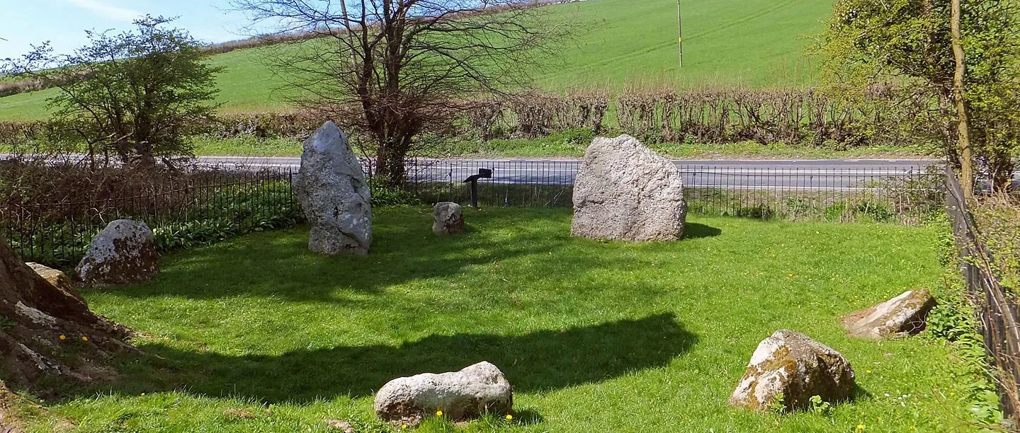 The Nine Stones, a small circle of grey standing stones on a green lawn inside iron railings, with a road and green fields beyond