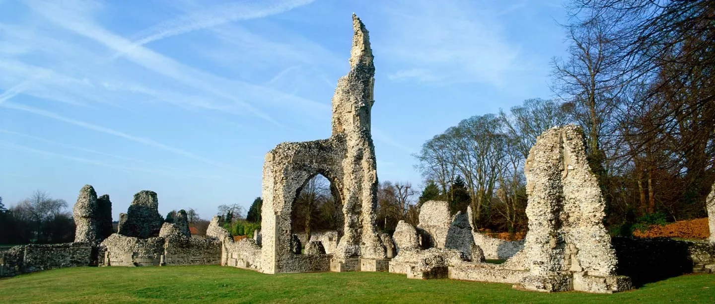 The ruined flint walls and tall arch of Thetford Priory on a sunlit lawn, with bare trees under a blue sky streaked with contrails