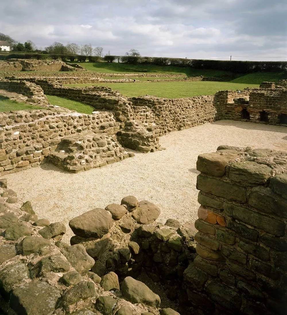 The excavated rooms of the Roman bath house at Wall, with sandstone and tile walls around gravelled floors and lawns beyond