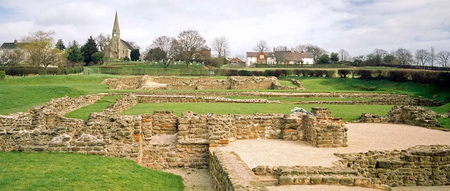 Low Roman stone walls of the bath house and mansio at Wall on a grassy slope, with the village church spire and houses beyond