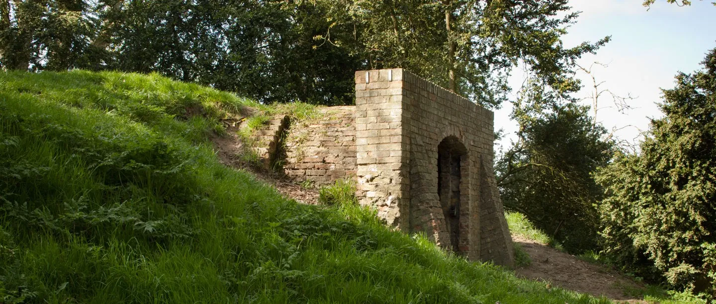 The brick ice house at Weeting Castle with a round-arched doorway, set into a grassy bank beneath trees in dappled sunlight