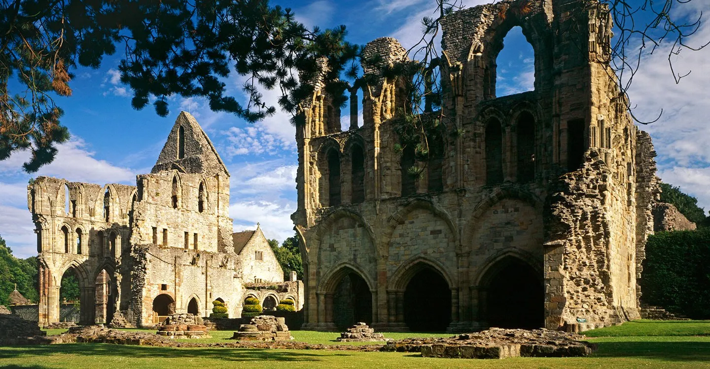 The ruined chapter house and south transept of Wenlock Priory framed by pine branches under a blue sky
