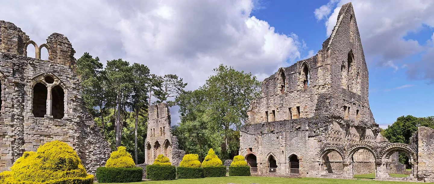 The ruined nave and transept of Wenlock Priory beyond rounded topiary yews under a stormy sky