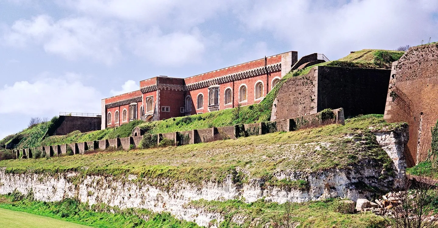 The brick barrack block of Western Heights above grassy ramparts, a loopholed wall and a white chalk ditch, under a cloudy sky
