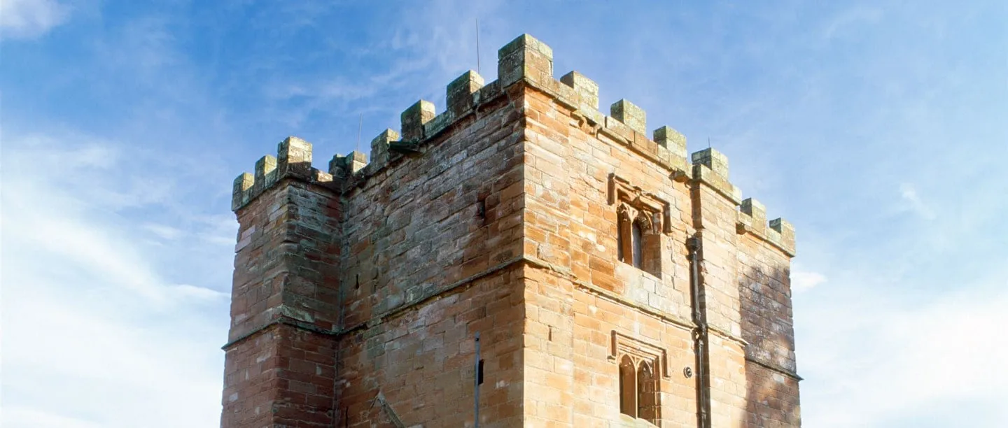 The battlemented red sandstone tower of Wetheral Priory Gatehouse with two Gothic windows, against a blue sky