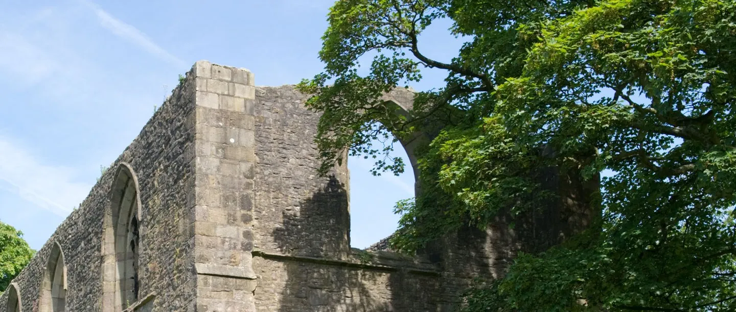 The ruined stone walls and empty arched windows of Whalley Abbey Gatehouse beside a sycamore in summer leaf