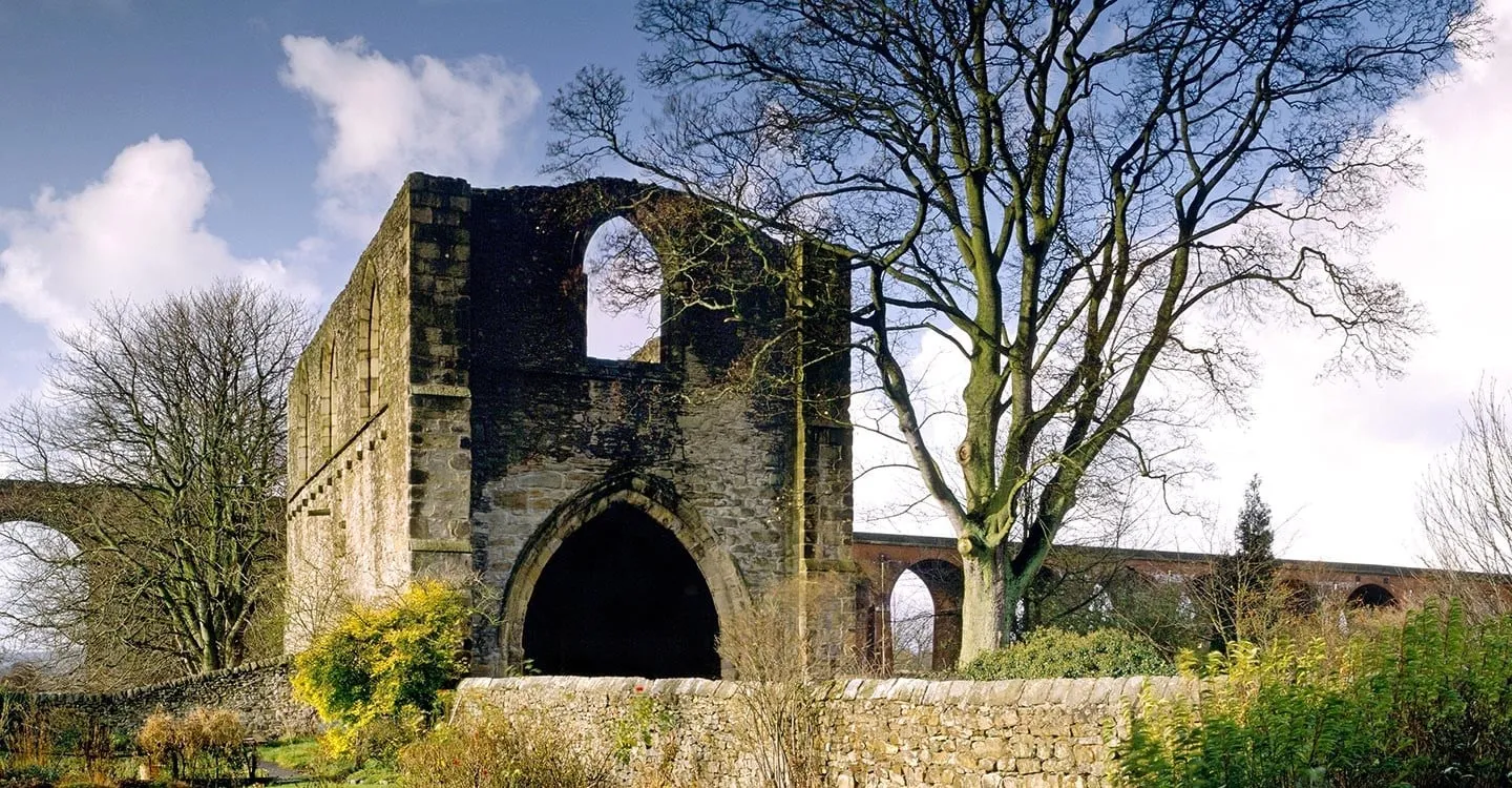 Whalley Abbey Gatehouse, a roofless stone gatehouse with a deep pointed arch, beside a bare tree with a brick railway viaduct behind