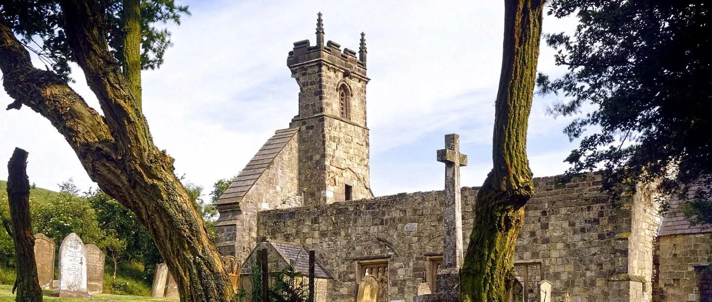 The ruined church of St Martin at Wharram Percy with its tower, gravestones and a churchyard cross, framed by gnarled trees